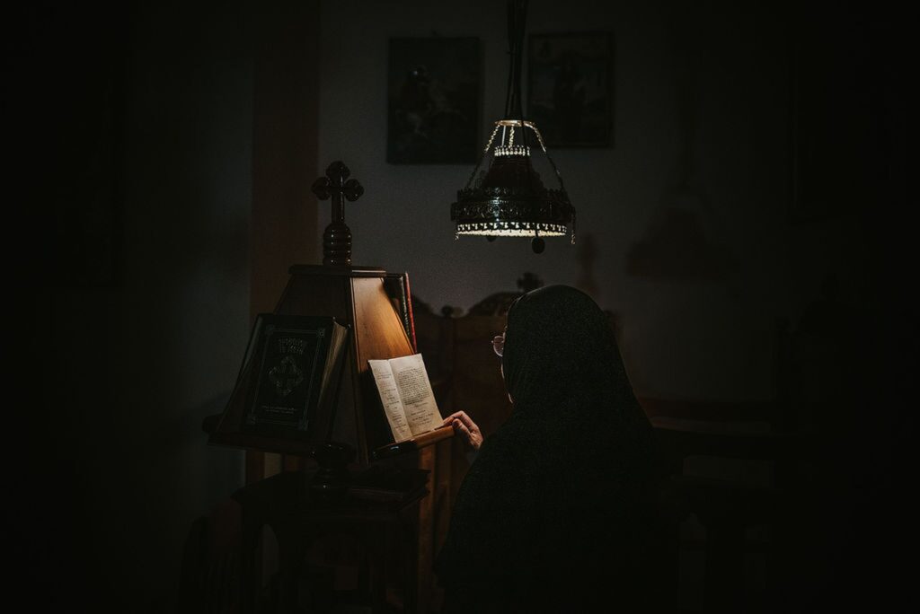 Person in dark religious attire reads from a book at a lectern under a dim hanging lamp | Esthesis Events Person in dark religious attire reads from a book at a lectern under a dim hanging lamp | Esthesis Events