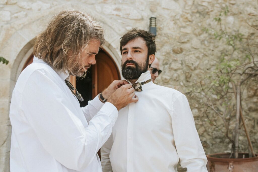 A groomsman helping Michael tie his bow tie before the wedding. | Esthesis Events A groomsman helping Michael tie his bow tie before the wedding. | Esthesis Events