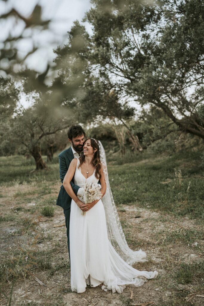 The bride and groom posing under the olive trees as Michael warmly embraces Jodie from behind. | Esthesis Events The bride and groom posing under the olive trees as Michael warmly embraces Jodie from behind. | Esthesis Events
